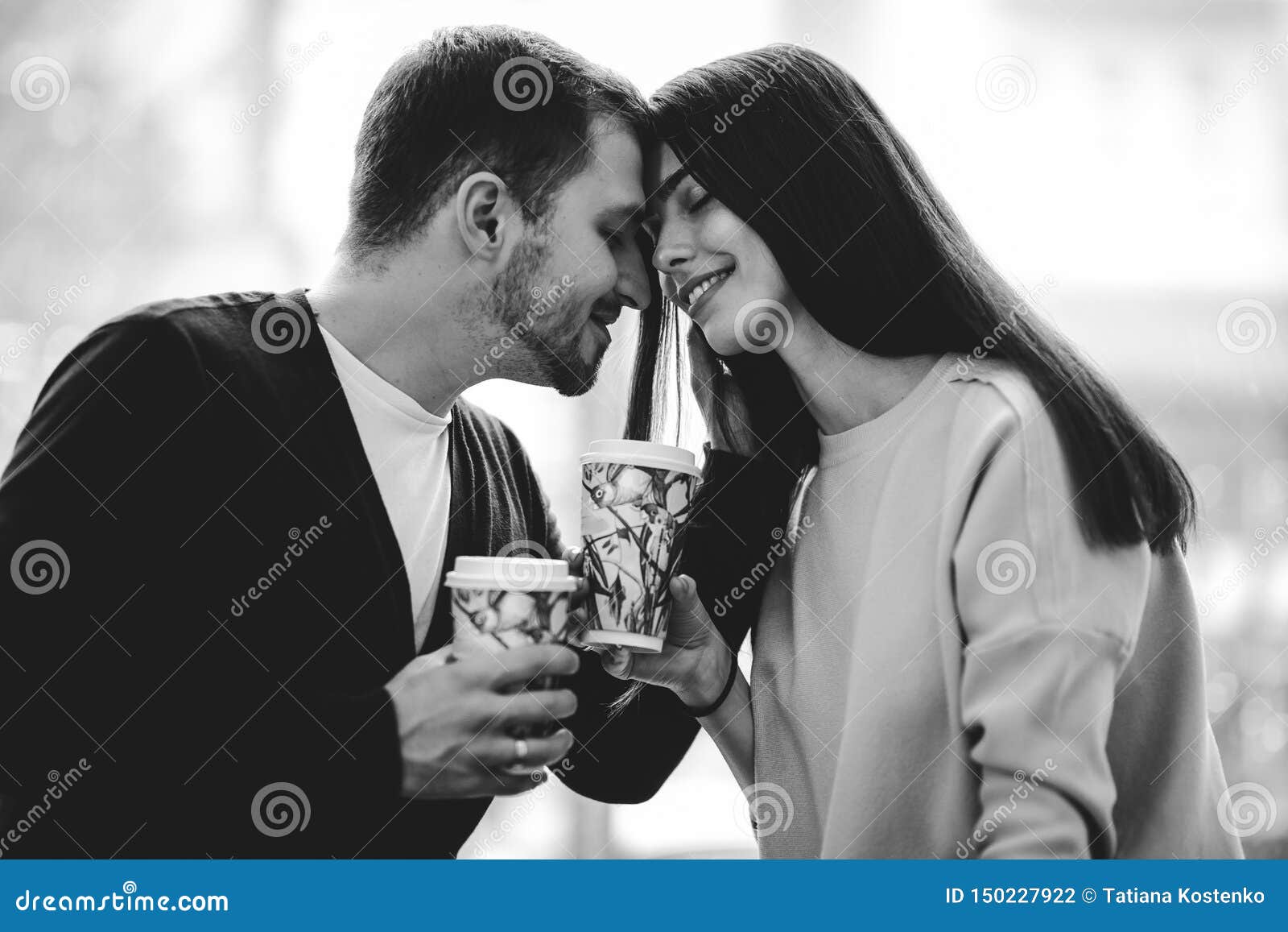 Loving Couple Is Siting Face To Face On The Background Of Window And Holding Cups In Their Hands