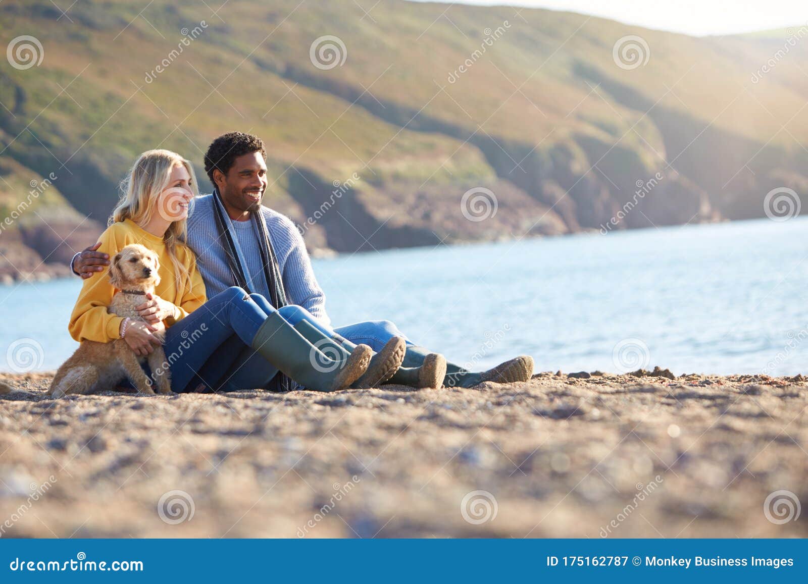 Loving Couple Sitting On Sand As They Walk With Dog Along Shoreline On Winter Beach Vacation