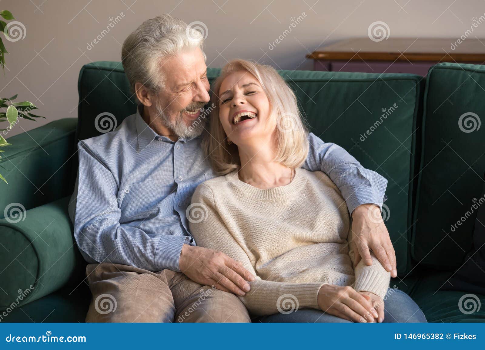 loving middle aged husband embracing laughing wife, sitting on couch