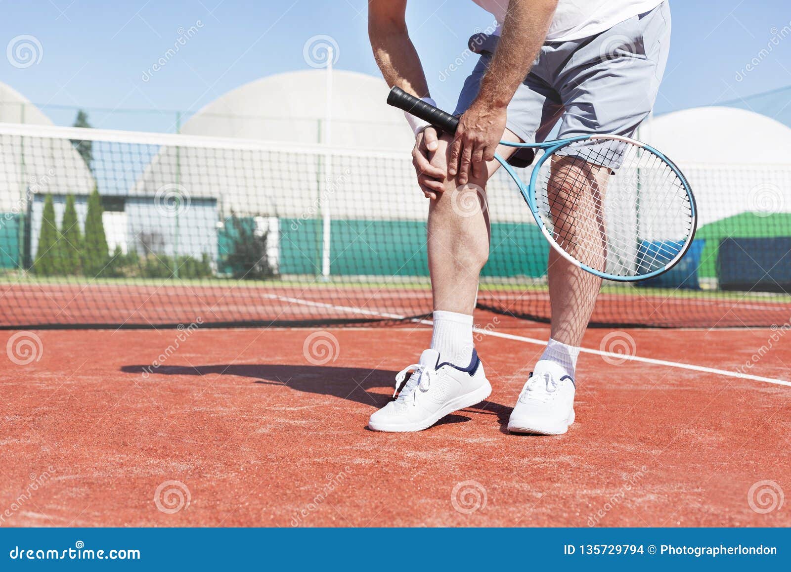 low section of mature man holding tennis racket while suffering from knee pain on red tennis court during summer