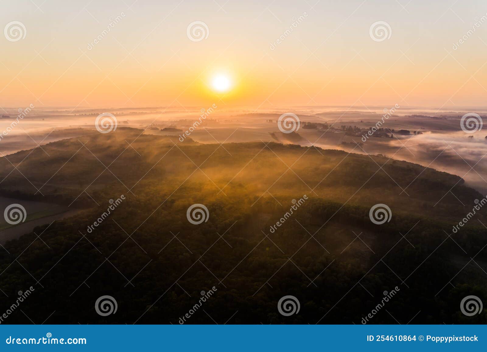 lush green forest surrounded by farm fields covered by fog with bright orange rising sun. birseye view of roztocze