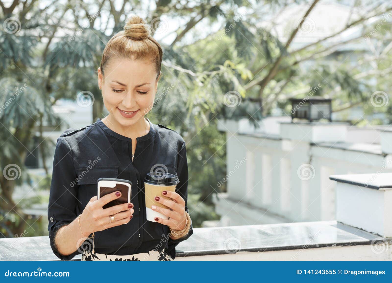 Lächelnde Geschäftsfrau mit Kaffee und Telefon. Positive hübsche Geschäftsfrau, die draußen mit Schale Mitnehmerkaffee steht und Textnachrichten liest