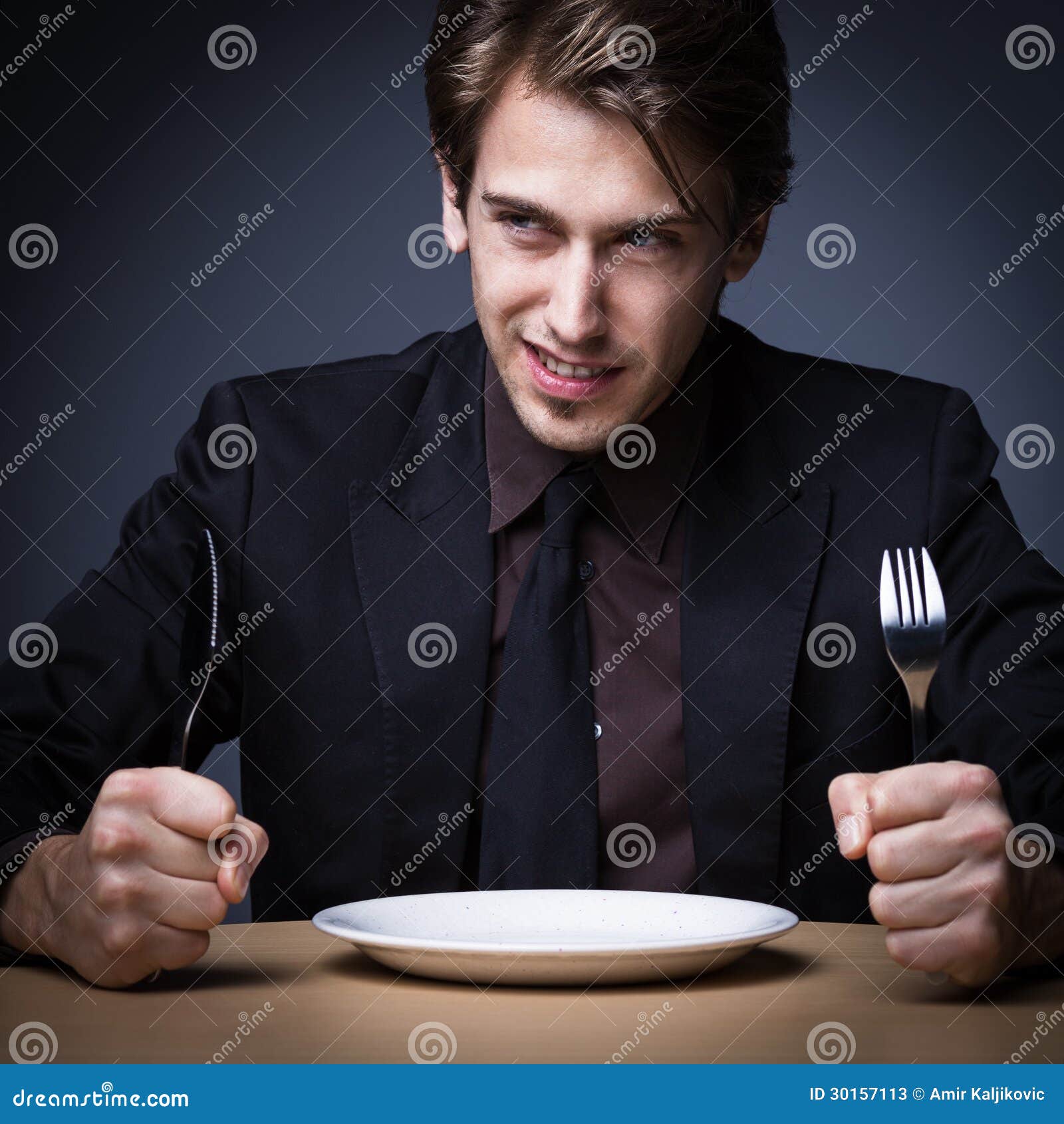Hungriger junger Mann. Lächelnder junger Mann, der Messer und Gabel aber mit leerer Platte auf der Tabelle mit dunkelgrauem Hintergrund hält.