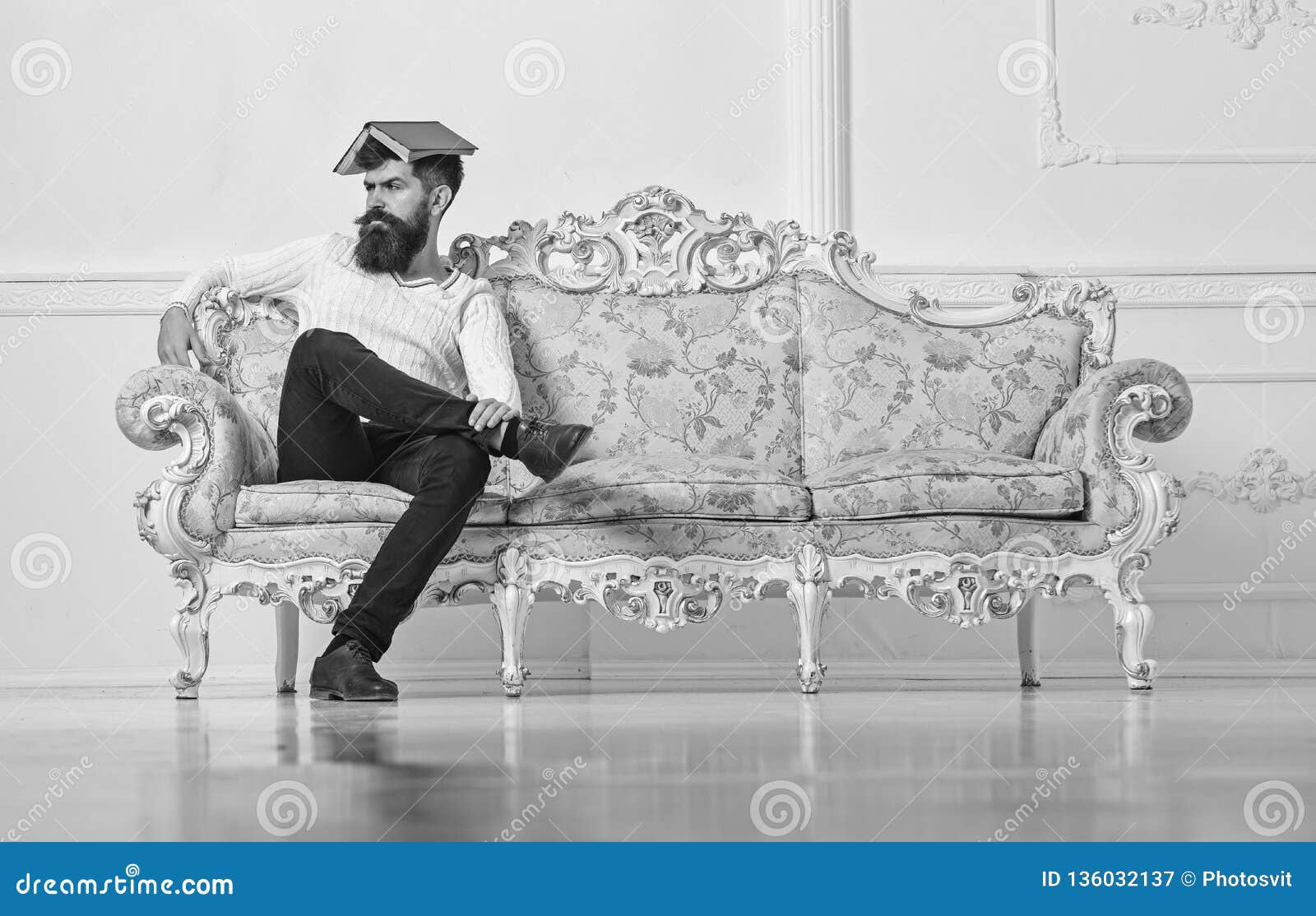 macho sits with open book on head, like roof. man with beard and mustache sits on sofa, white wall background. overstudy