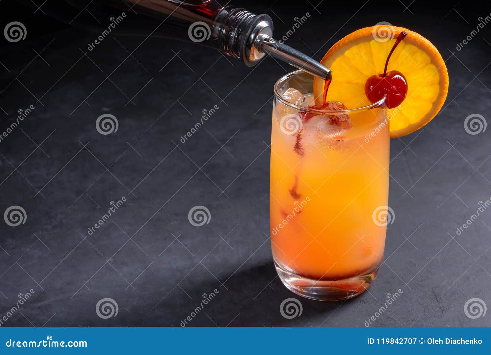 making tequila sunrise cocktail. grenadine slowly poured into a glass of ice, orange juice and tequila. dark stone background with