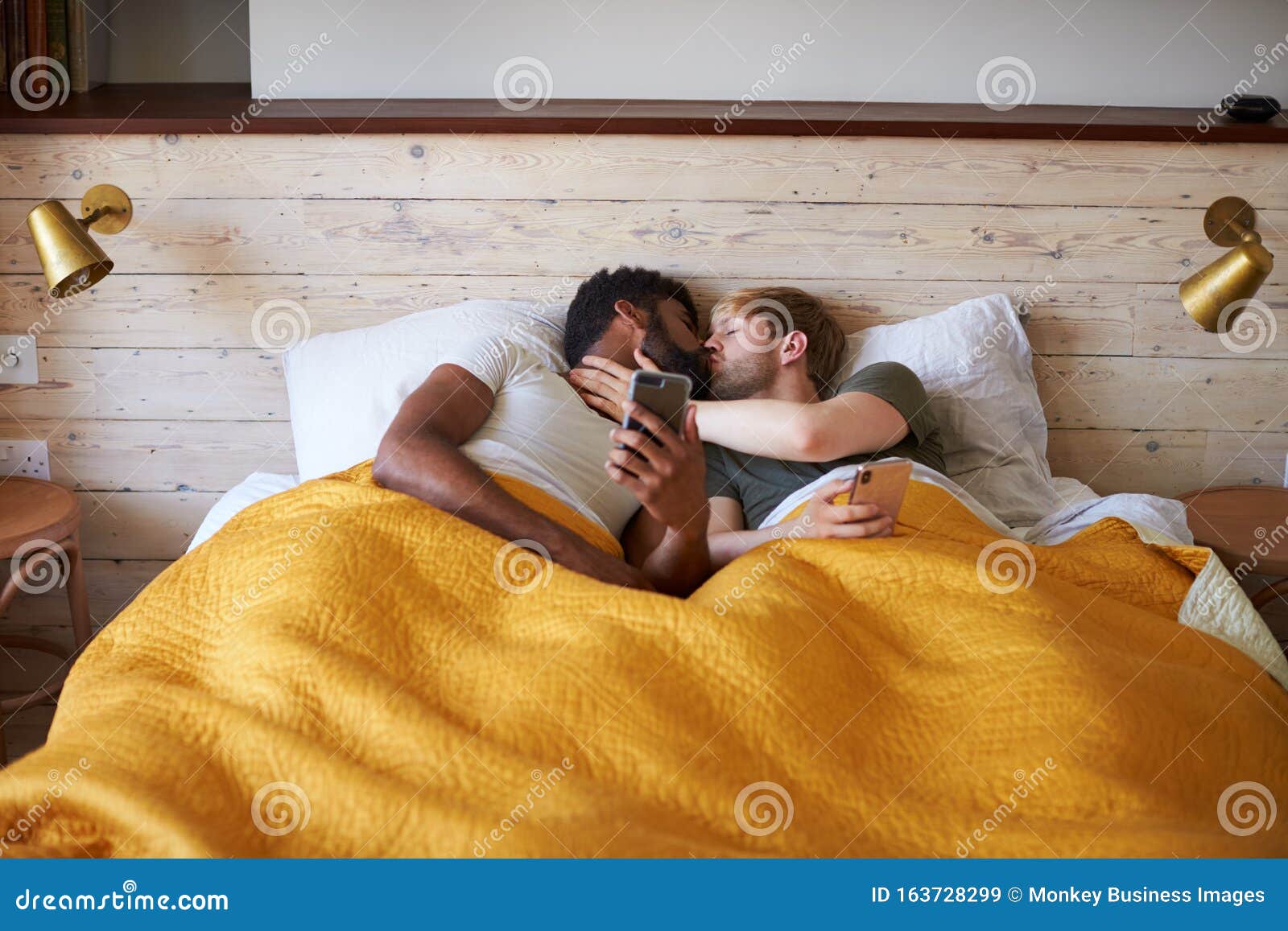 Male Gay Couple Lying In Bed At Home Checking Mobile
