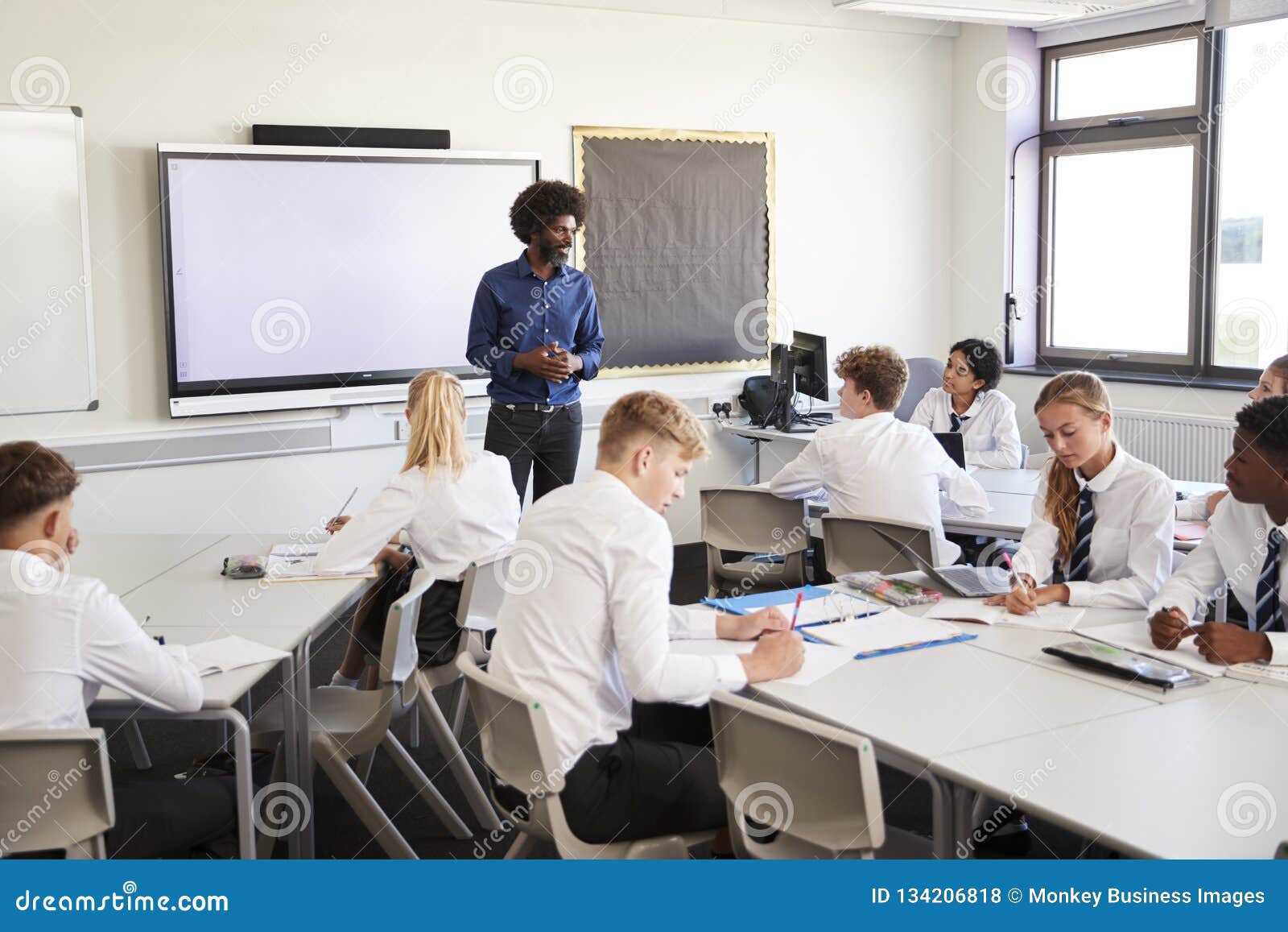 male high school teacher standing next to interactive whiteboard and teaching lesson to pupils wearing uniform