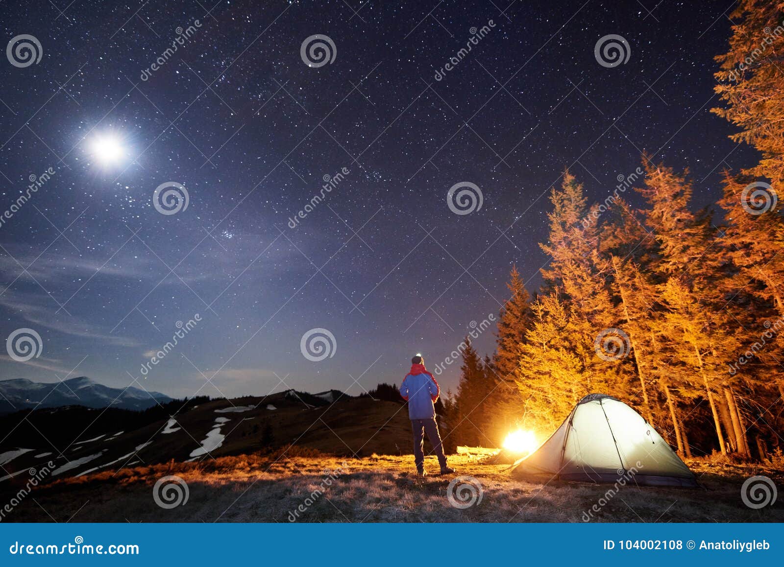 male hiker have a rest in his camp near the forest at night under beautiful night sky full of stars and the moon