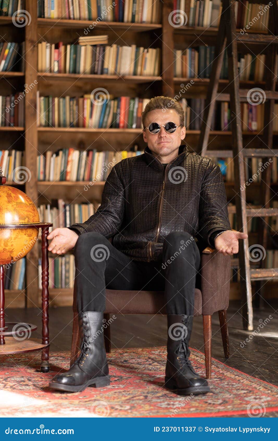 Male In Hipster Leather Jacket And Stylish Sunglasses Sits In A Retro Armchair With Open Hands