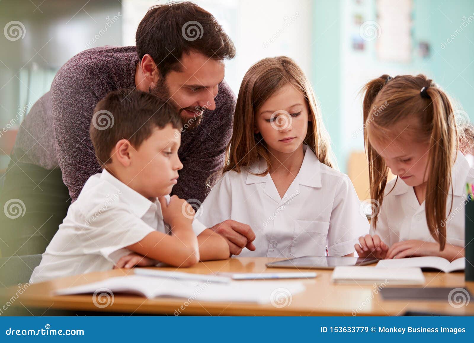 Male Teacher with Three Elementary School Pupils Wearing Uniform Using Digital Tablet at Desk