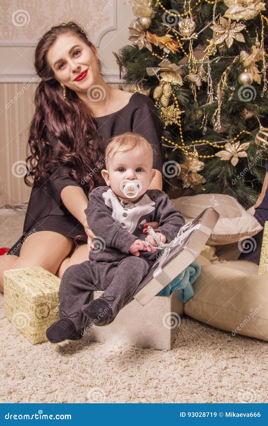 Mamman sitter under trädet med ett småbarn i en gåvaask. Den lyckliga mamman sitter under en julgran med ett småbarn i en gåvaask