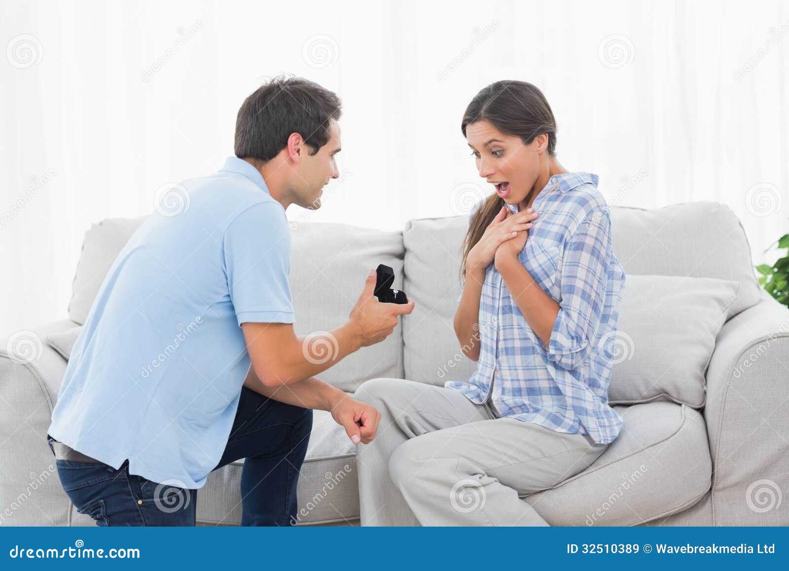 Man on bended knee offering an engagement ring to his partner on the couch