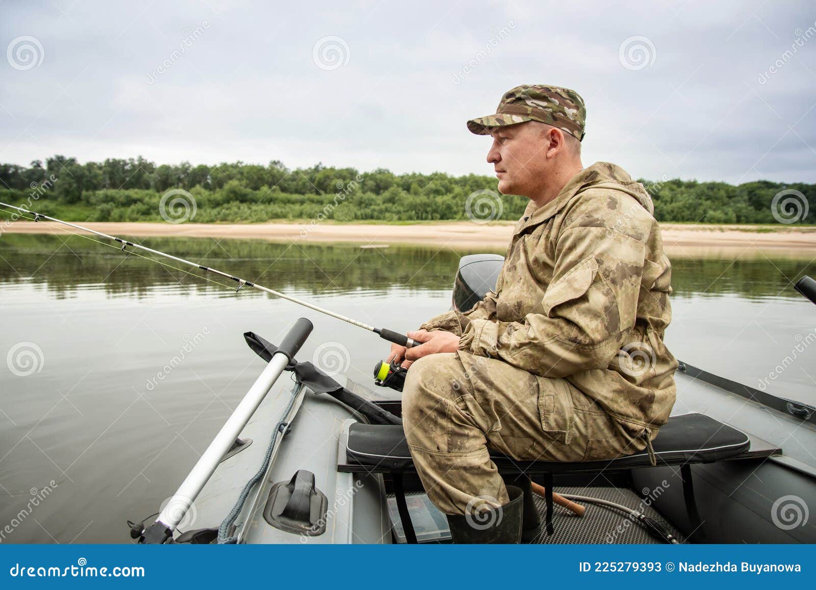 A Man in a Camouflage Suit and Cap Sits in a Boat with a Fishing