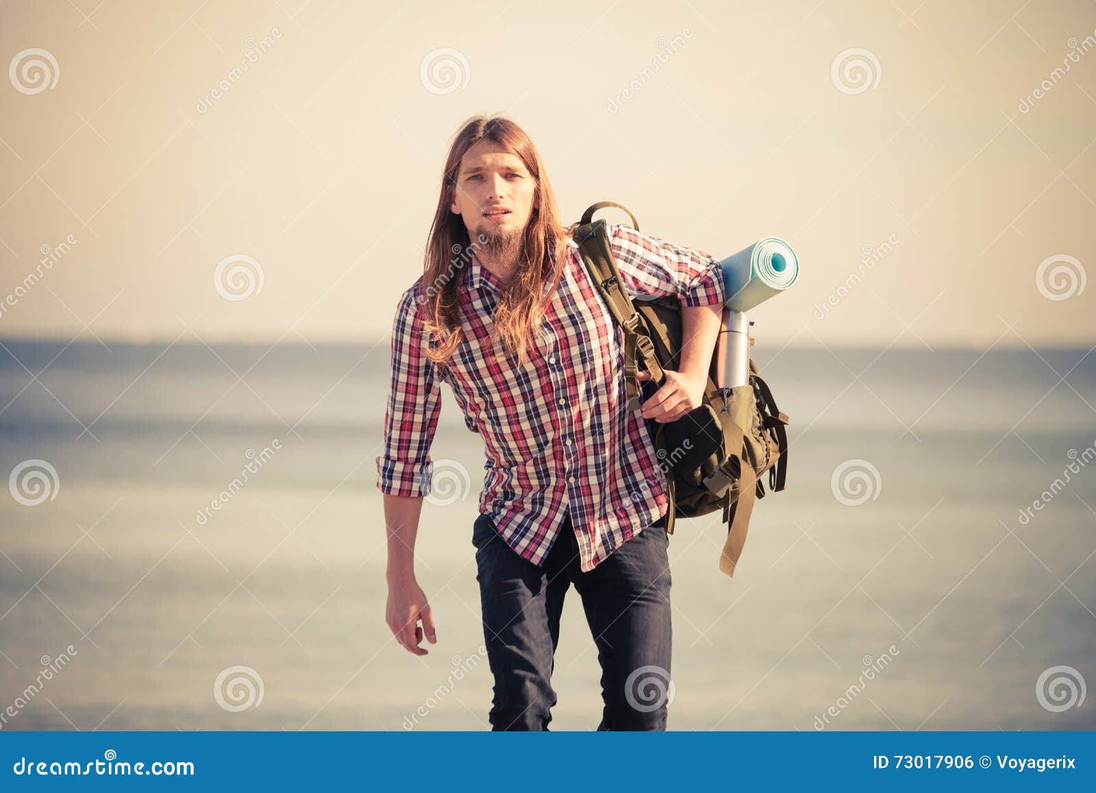 Man hiker with backpack tramping by seaside. Man hiker backpacker walking with backpack by seaside at sunny day. Adventure, summer, tourism active lifestyle. Young long haired guy tramping