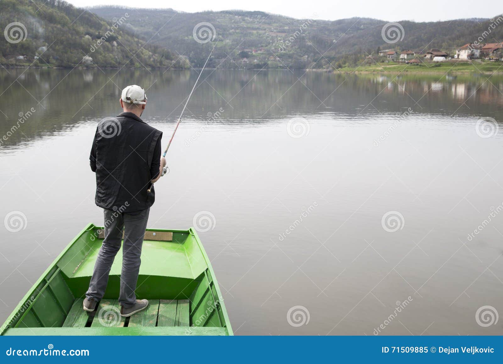 man with his back towards camera fishing from the boat reeling string and waiting fish to take a bait water reflection