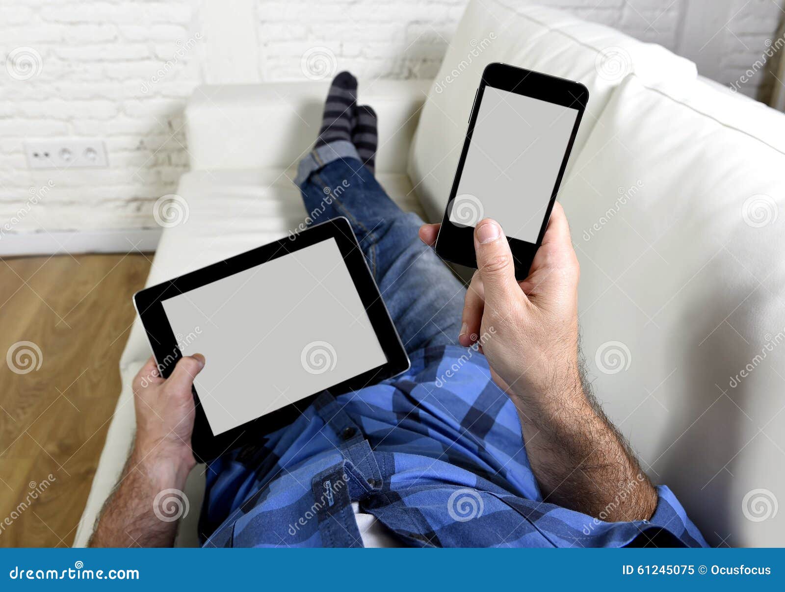 man lying on couch at home living room using simultaneously mobile phone and digital tablet pad in internet technology