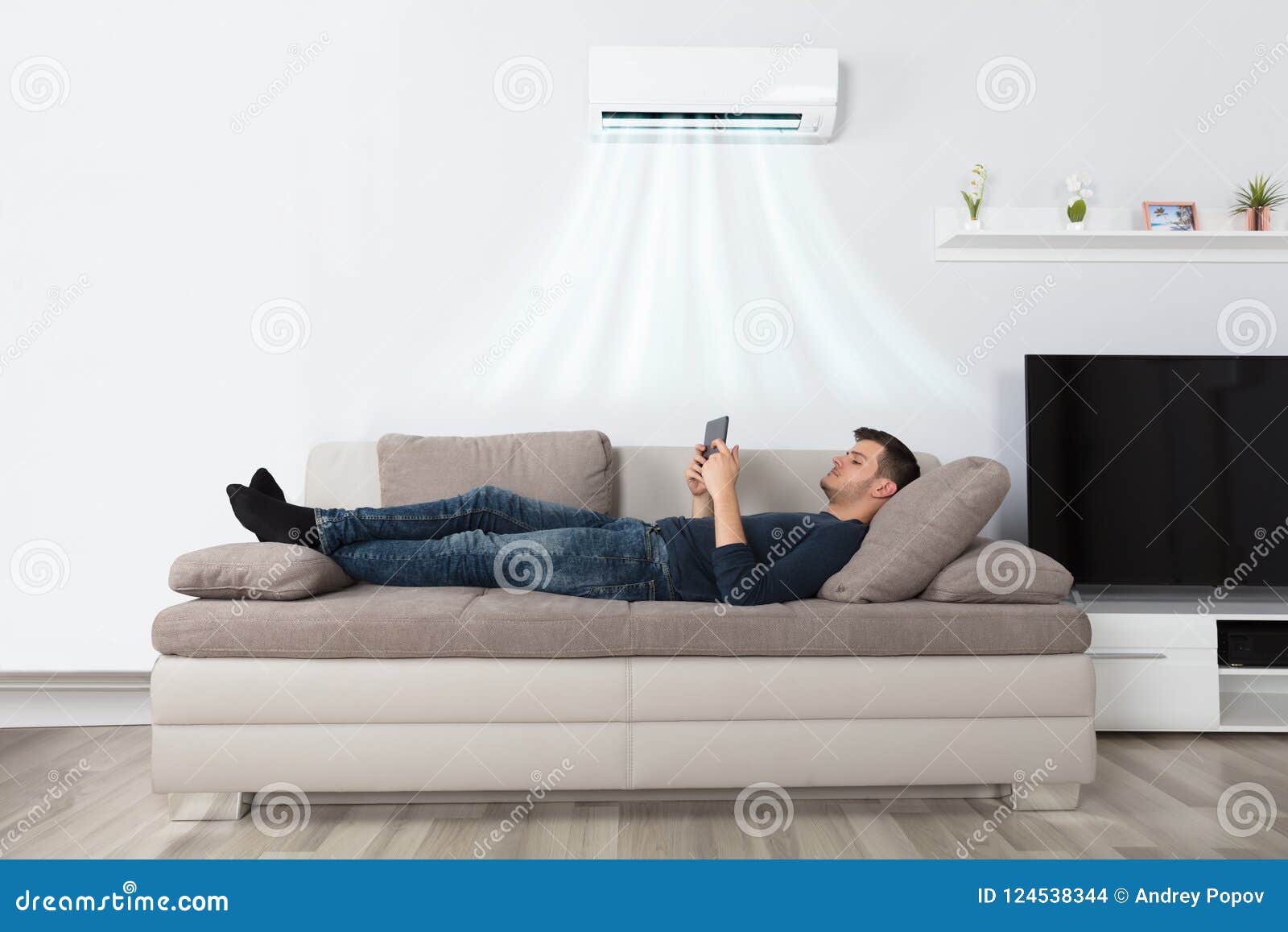 man lying on couch under air conditioner using tablet
