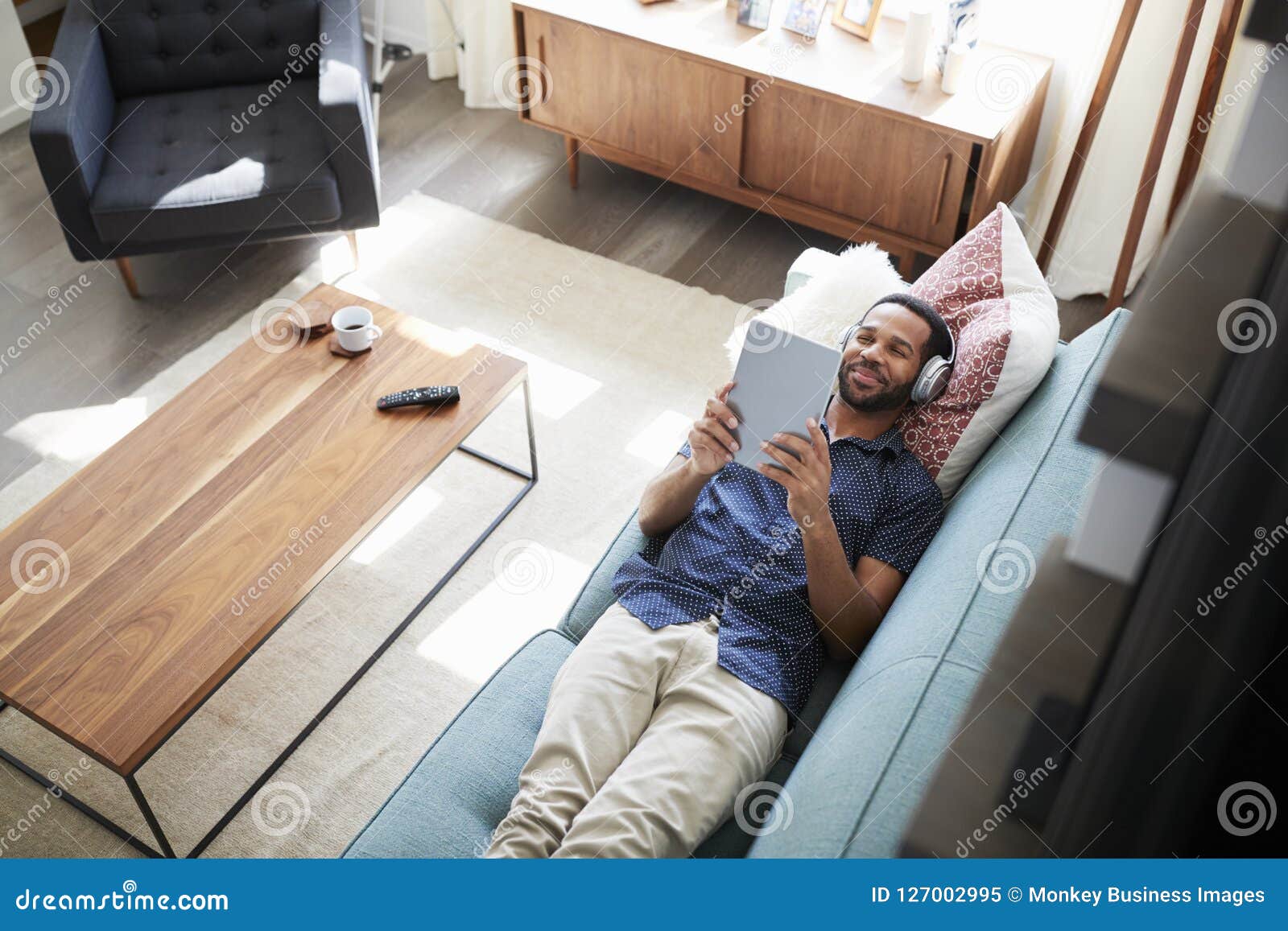 man lying on sofa at home wearing headphones and watching movie on digital tablet