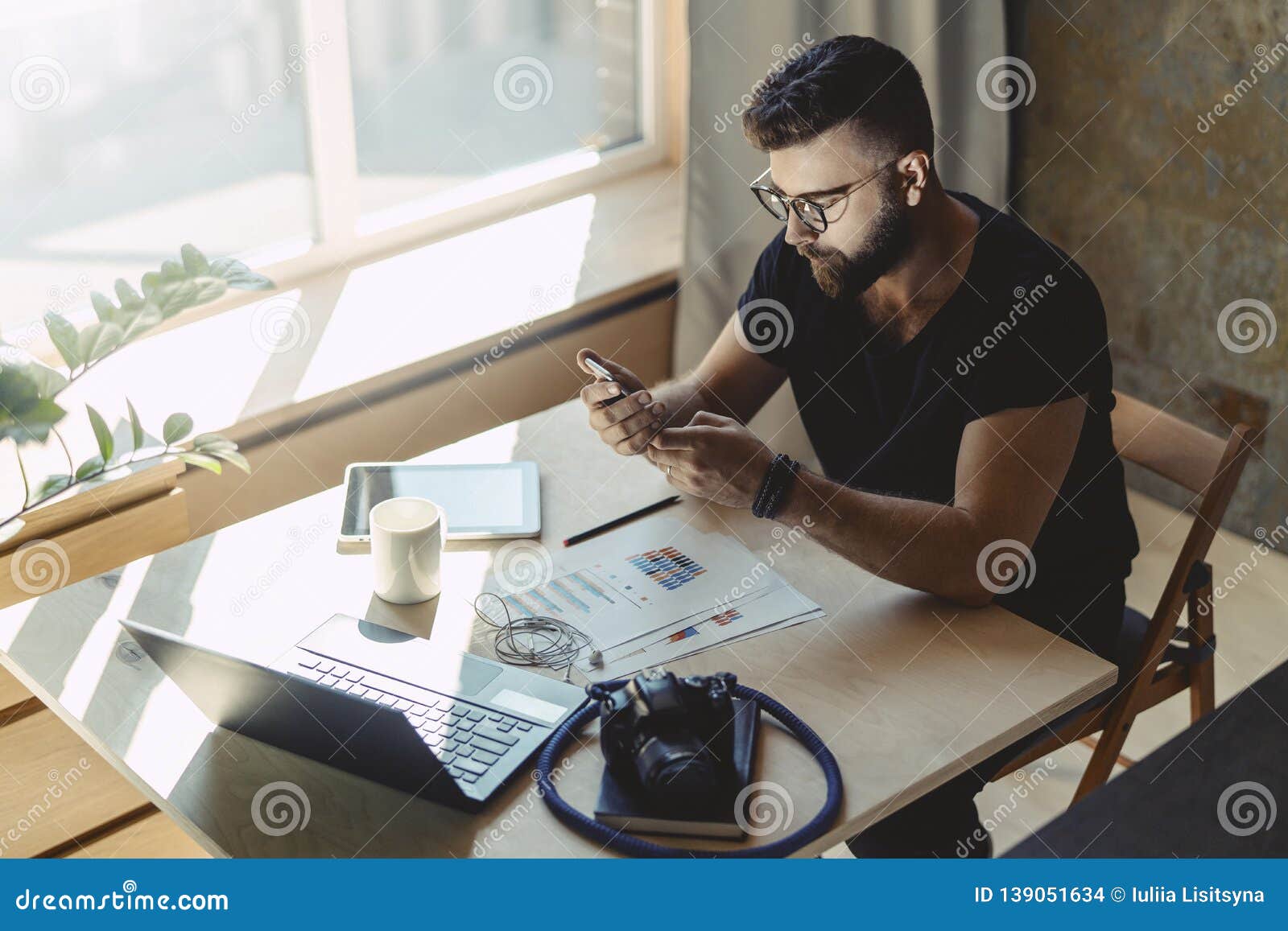 man millenial in glasses sits at table in front of laptop and uses smartphone, reads message from business partner