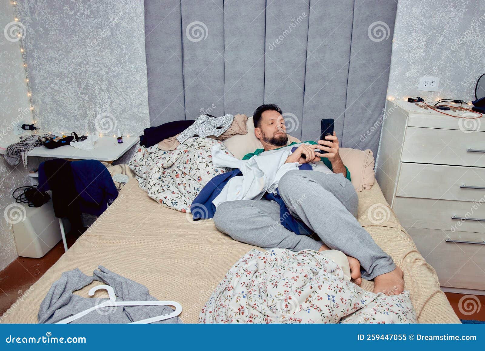 A Man with a Phone in His Hand is Lying in the Bedroom on the Bed , Scattered Clothes and a Mess