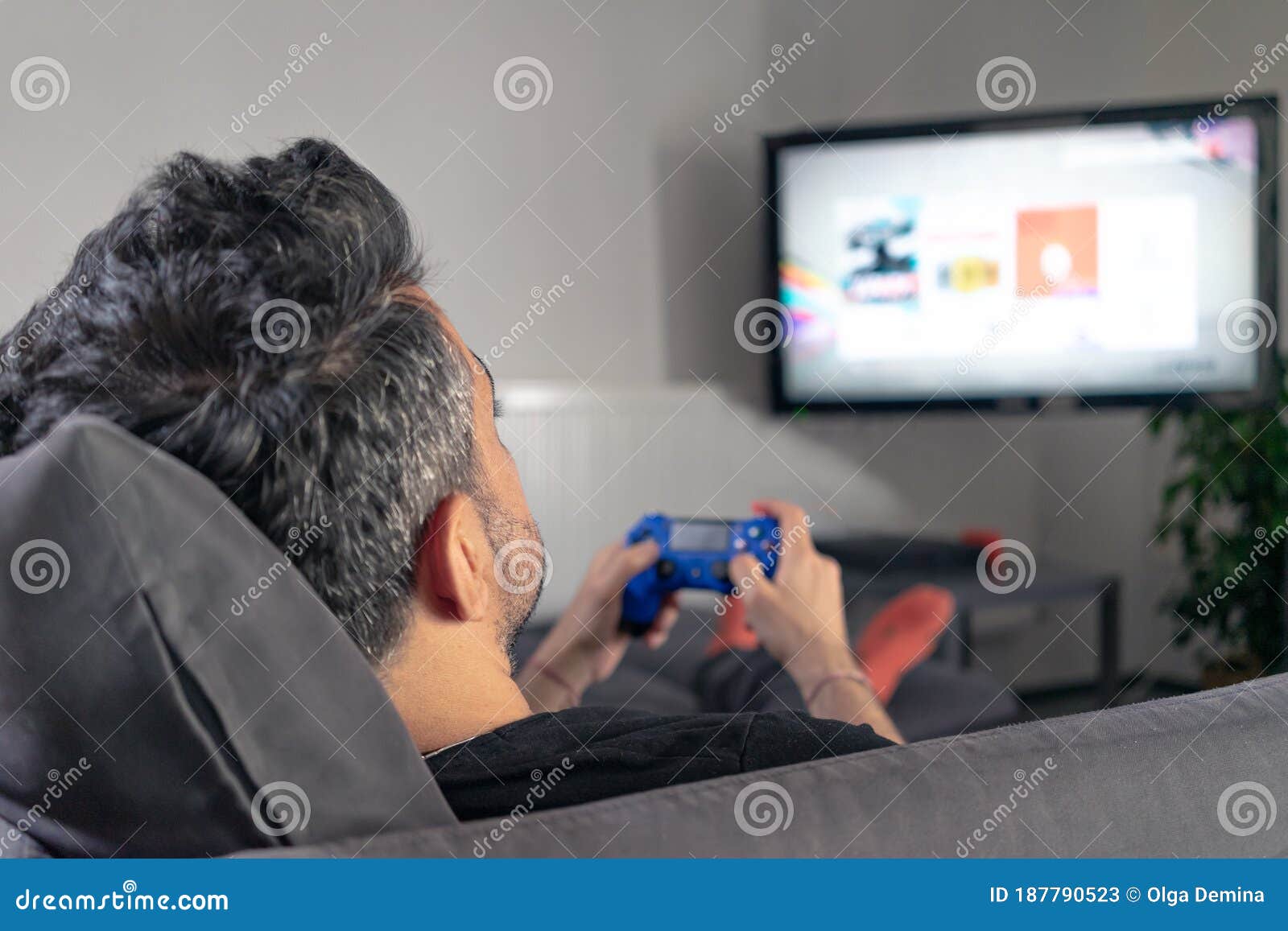 man playing video game at home sitting on coach sofa in front of television with play game concole and joystick in hands