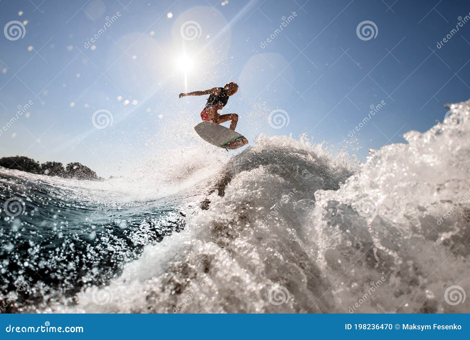 man professionally rides and bounces on surf style wakeboard over splashing wave against blue sky