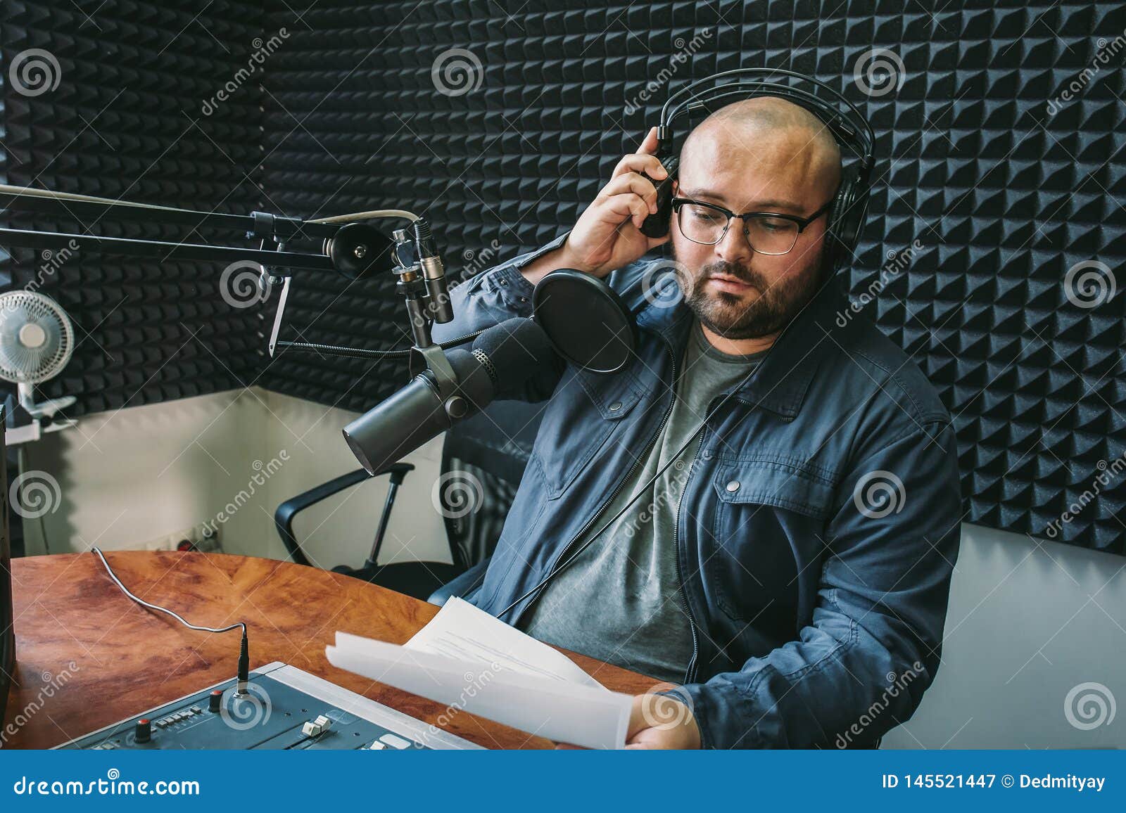 man radio host or representer or journalist reads news from paper list in hand to studio microphone sitting at mixing sound panel