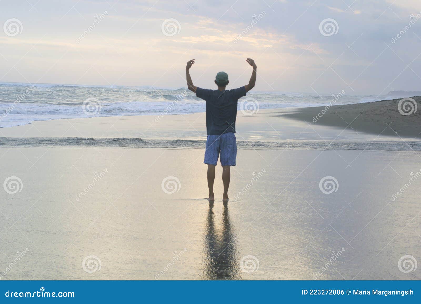 man standing alone from behind on the beach, with arm raised against the sea at sunset.