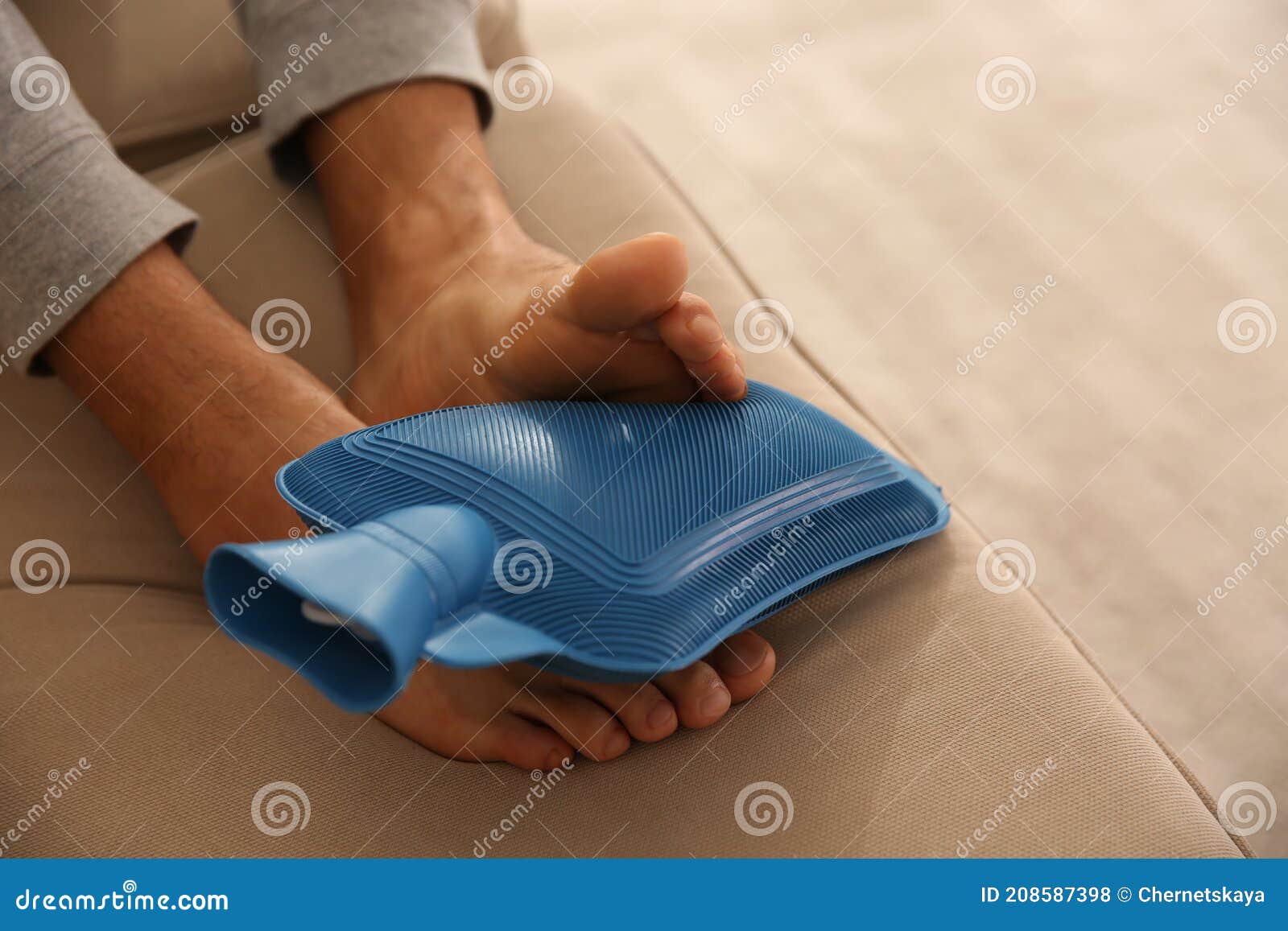 Man Warming Feet with Hot Water Bottle on Sofa, Closeup Stock Photo