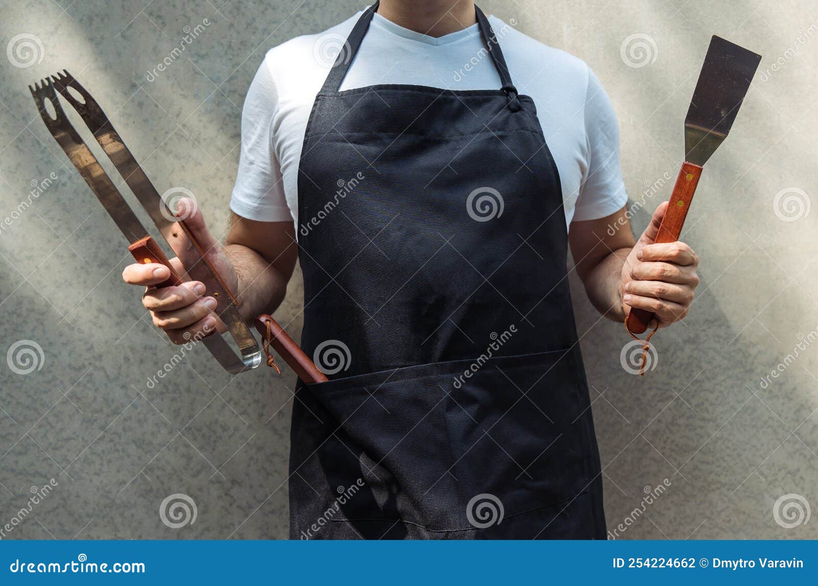 a man wearing black chef`s apron, holding barbecue tools: bbq tongs, spatula