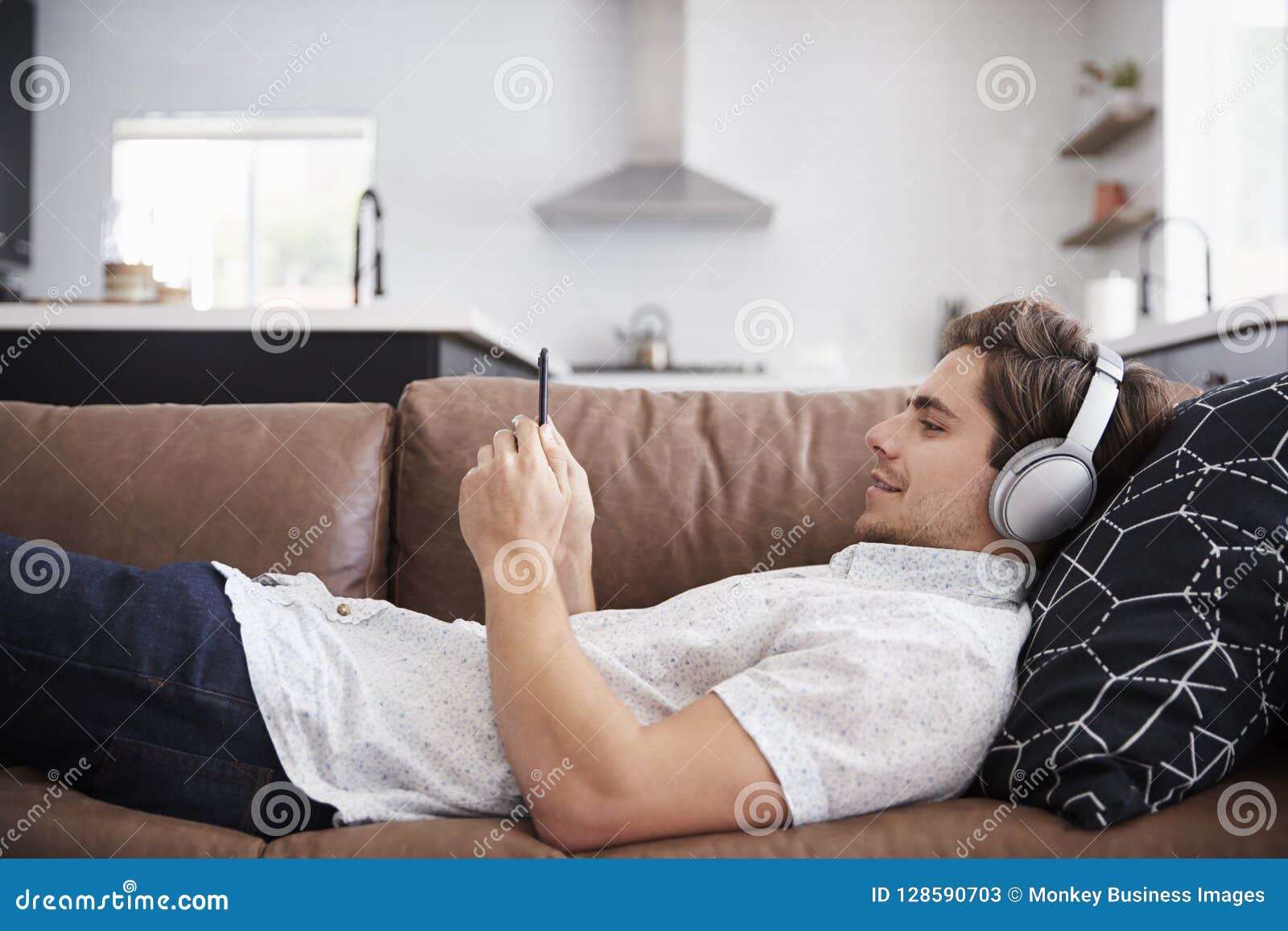 man wearing wireless headphones lying on sofa at home streaming from mobile phone