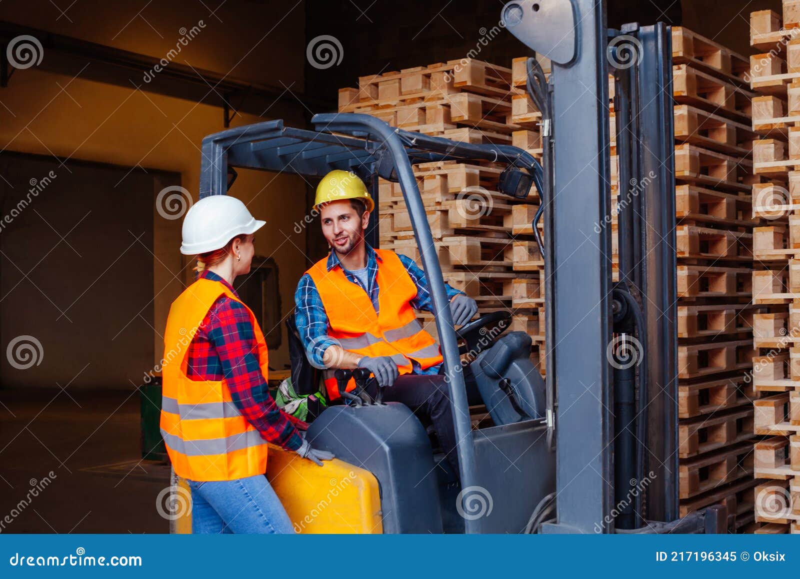 man worker sitting in industrial forklift at warehouse talking to woman co-worker