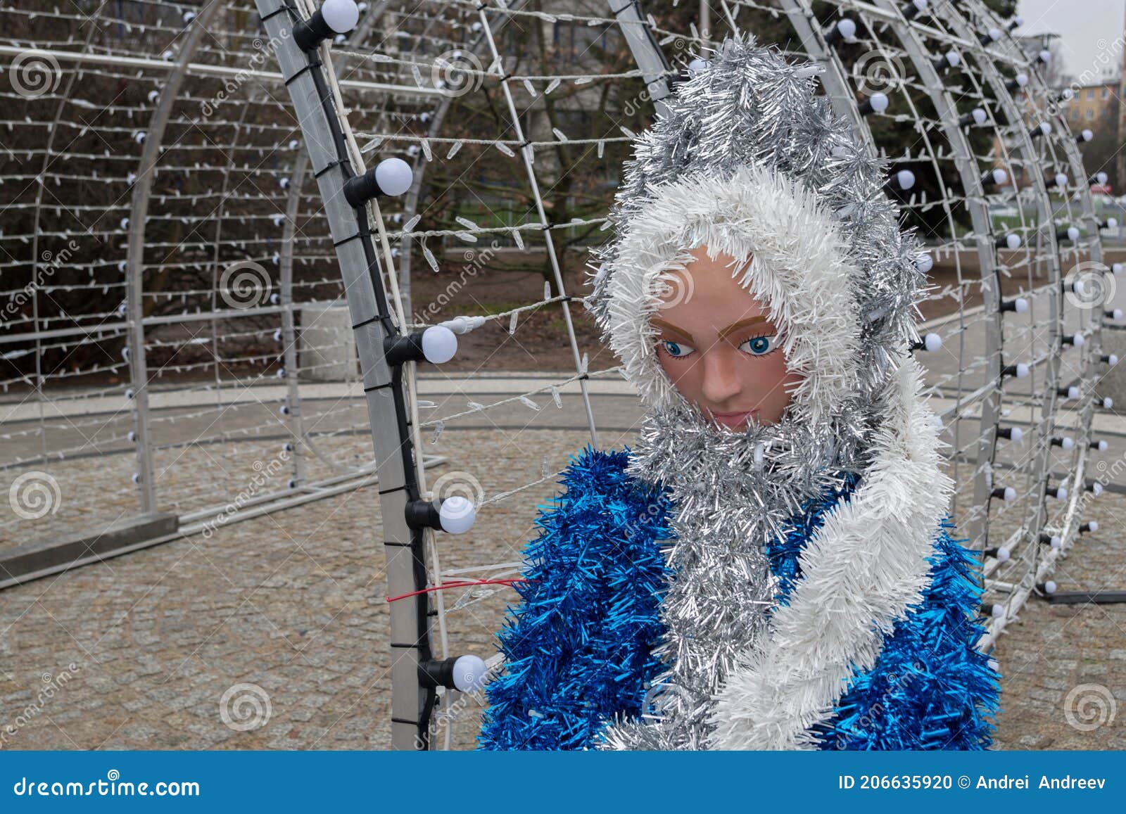 mannequin of snow maiden in blue fluffy fur coat with white fringe standing on stone platform near arch with rows of lamps