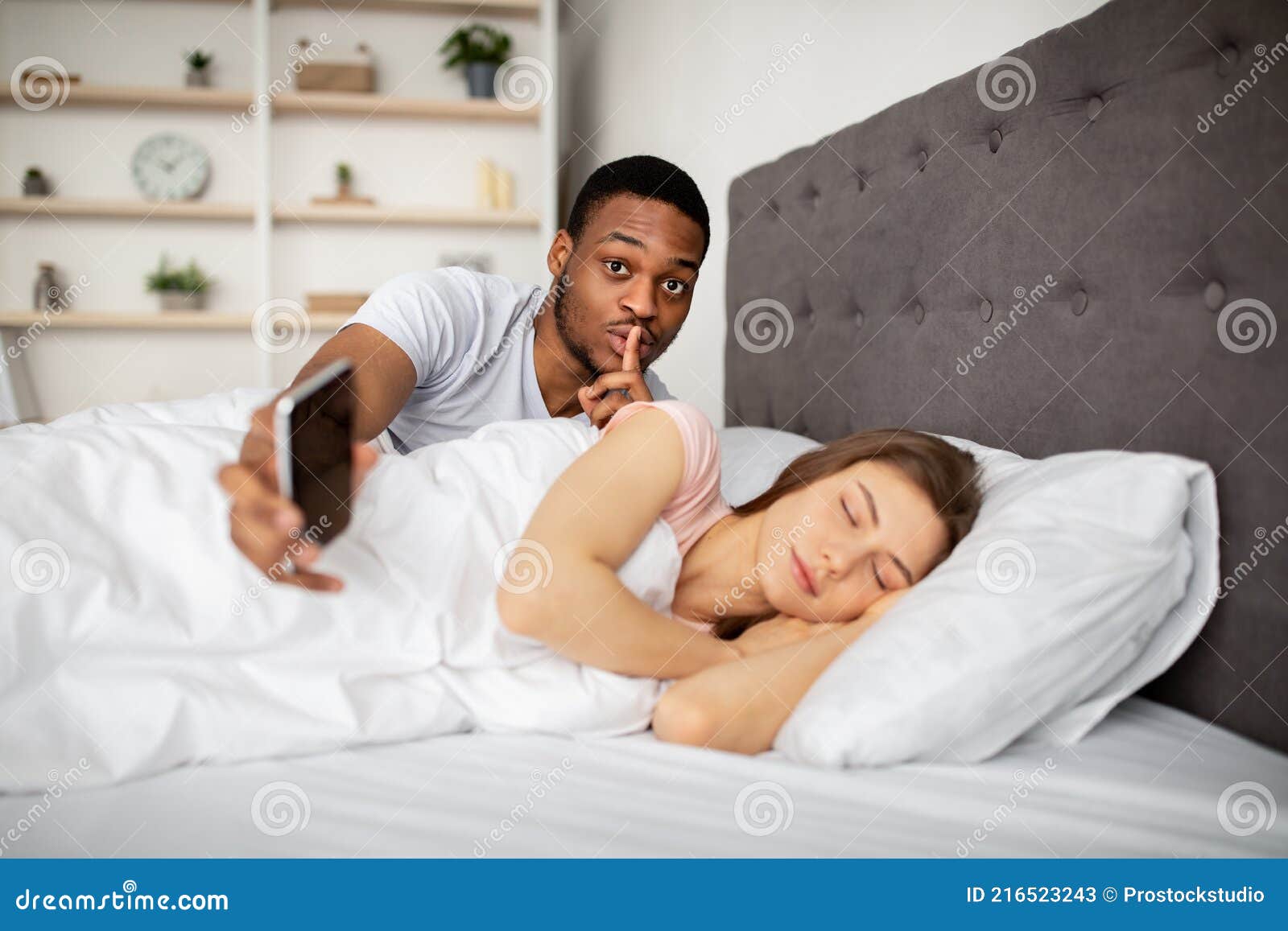 Marido Negro Ciumento Espiando Sua Esposa Mostrando Gesto De Silêncio Verificando Seu Celular Enquanto Ela Dorme Na Cama Imagem de Stock