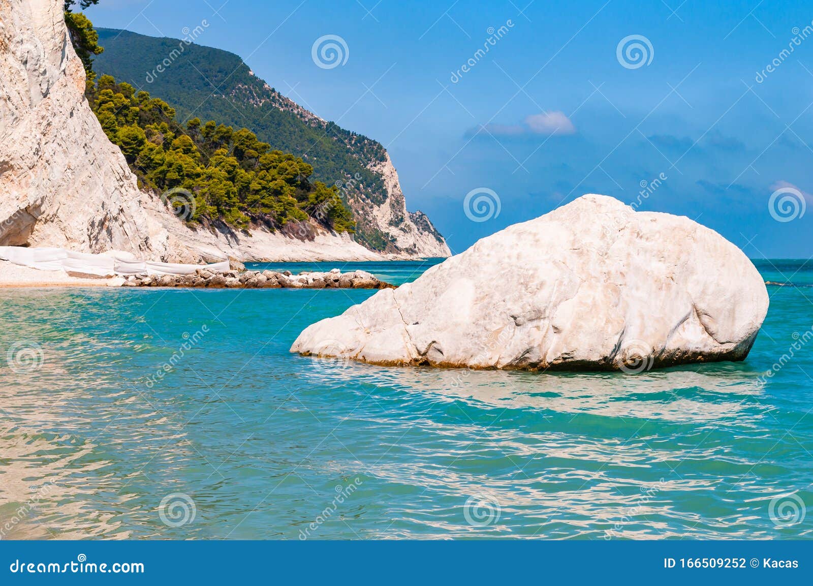 massive white boulder as a sea stack lying in water of adriatic sea with beautiful coastline of numana, italy on the background.