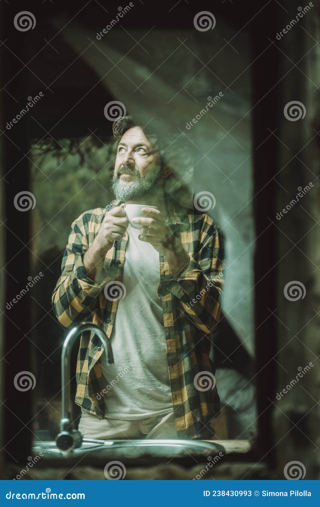 mature adult man at home viewed from outside thrugh the glass winws drinking coffee and looking around - alternative rural