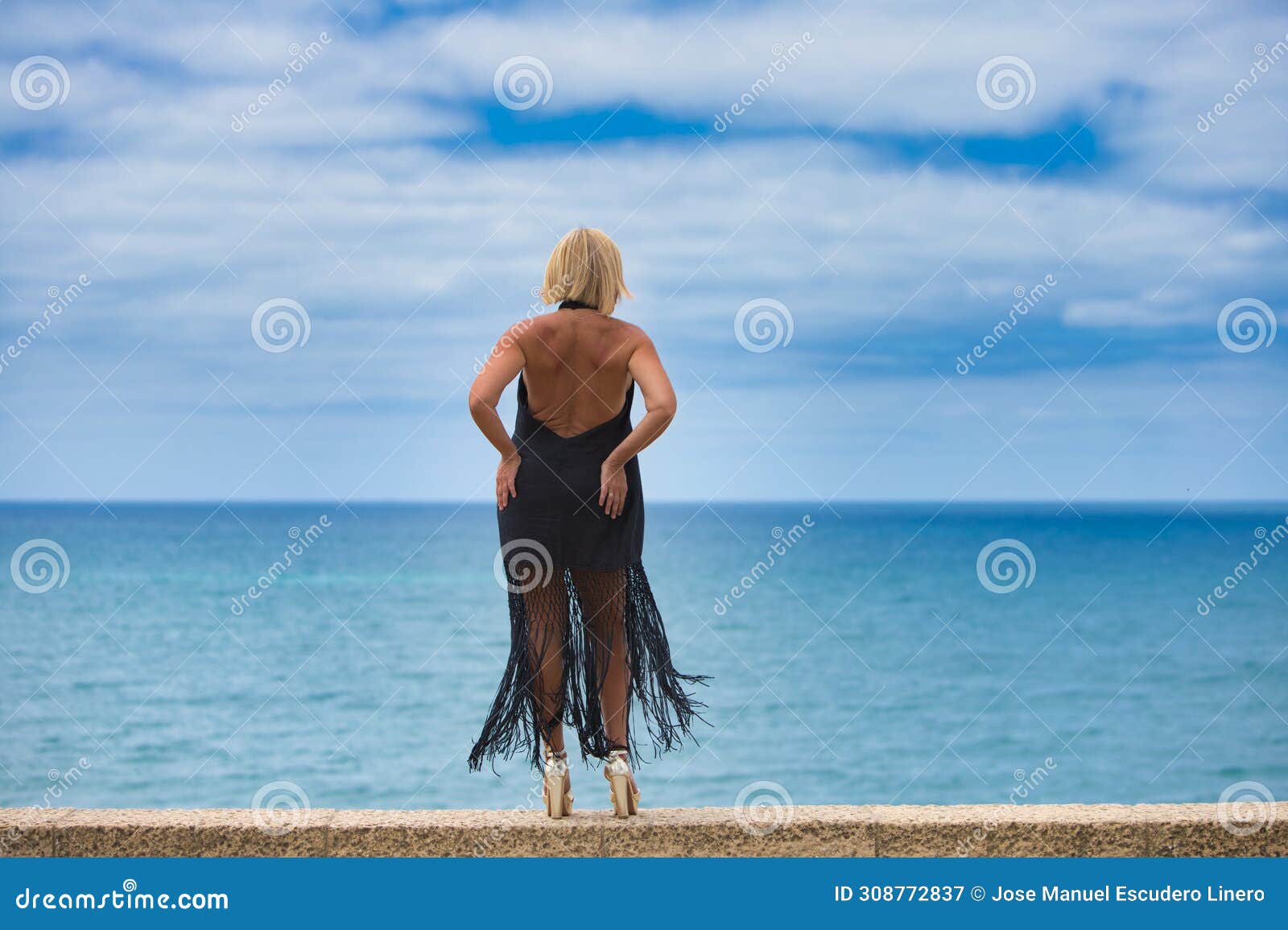 mature, blonde, beautiful woman wearing an elegant black dress gazes longingly at the horizon over the atlantic ocean in cadiz,