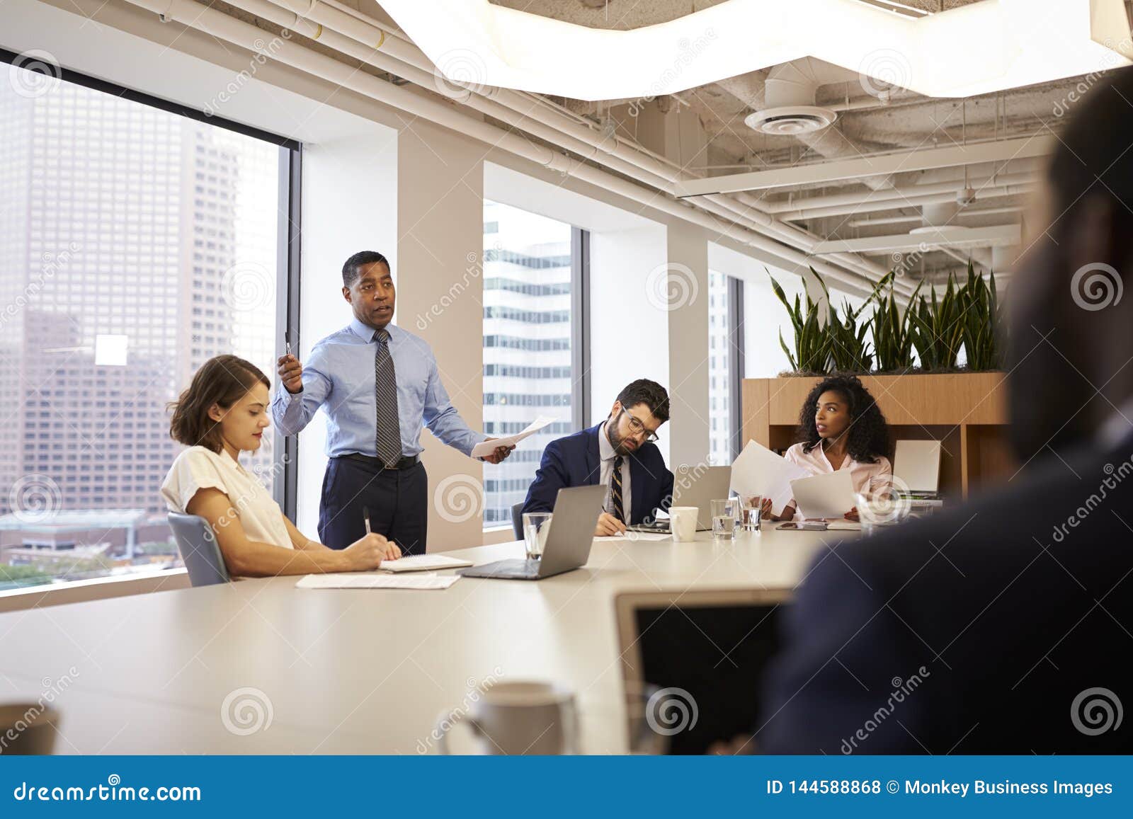 Mature Businessman Standing Giving Presentation To Colleagues In Modern Open Plan Office