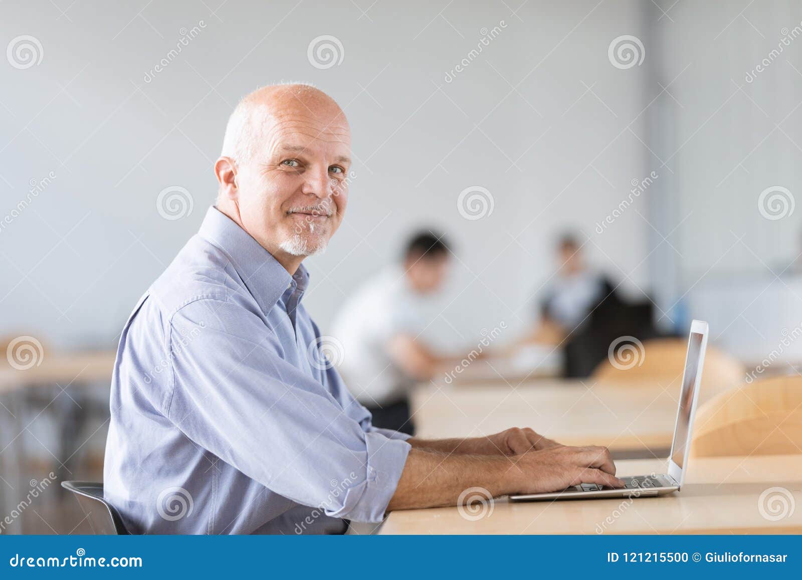 mature man seated and looking at camera while working on on lapt