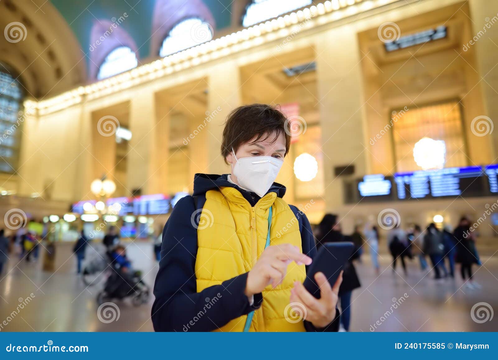 mature woman wearing a face mask is using her phone to buy a ticket online in grand central station new york, manhattan. female