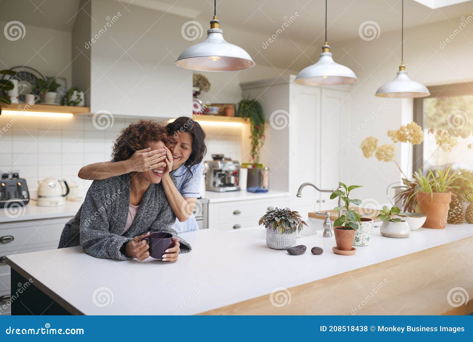 Mature Woman Wearing Pyjamas Surprising Same Sex Partner in Kitchen at Home Together Stock Photo