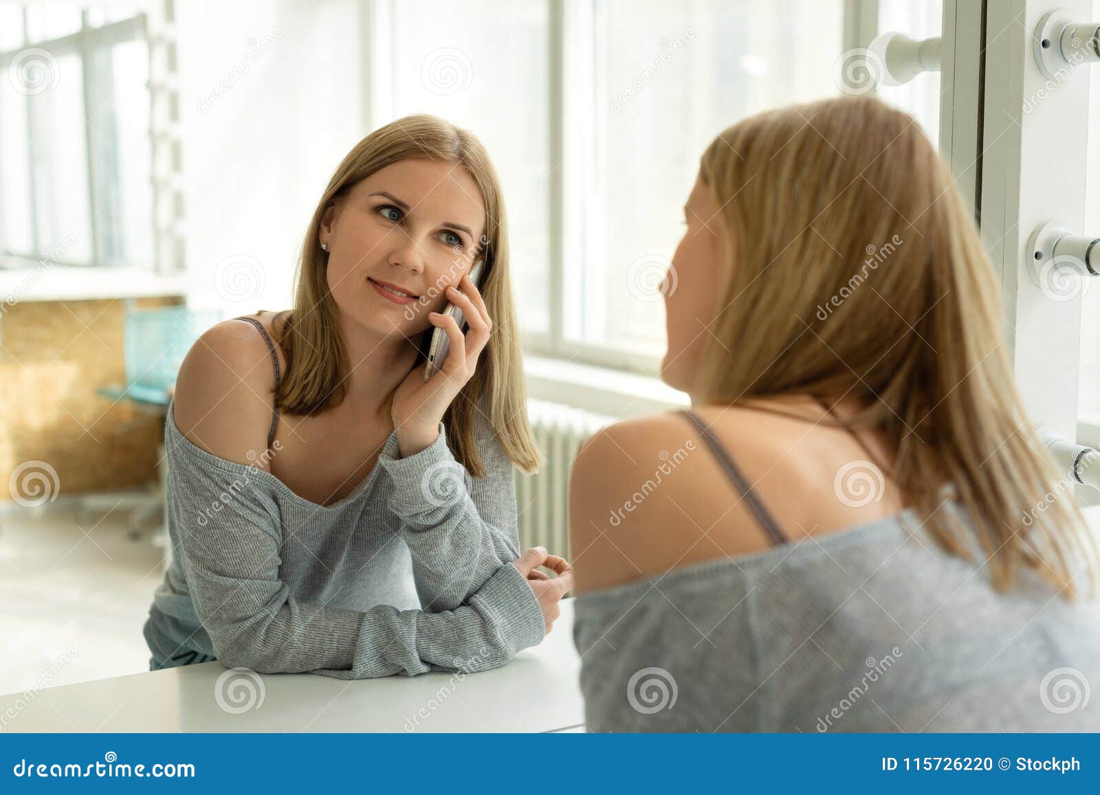 Menina consideravelmente loura dos jovens que fala no telefone na frente de um espelho em uma sala brilhante