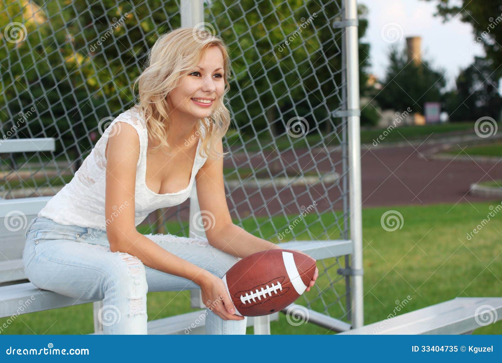 Menina loura feliz com futebol americano. Jovem mulher bonita alegre de sorriso que senta-se no banco. Fora. Fã do futebol. Menina loura feliz com futebol americano. Jovem mulher bonita alegre de sorriso que senta-se no banco. Fora. O fã da equipa de futebol está olhando o jogo