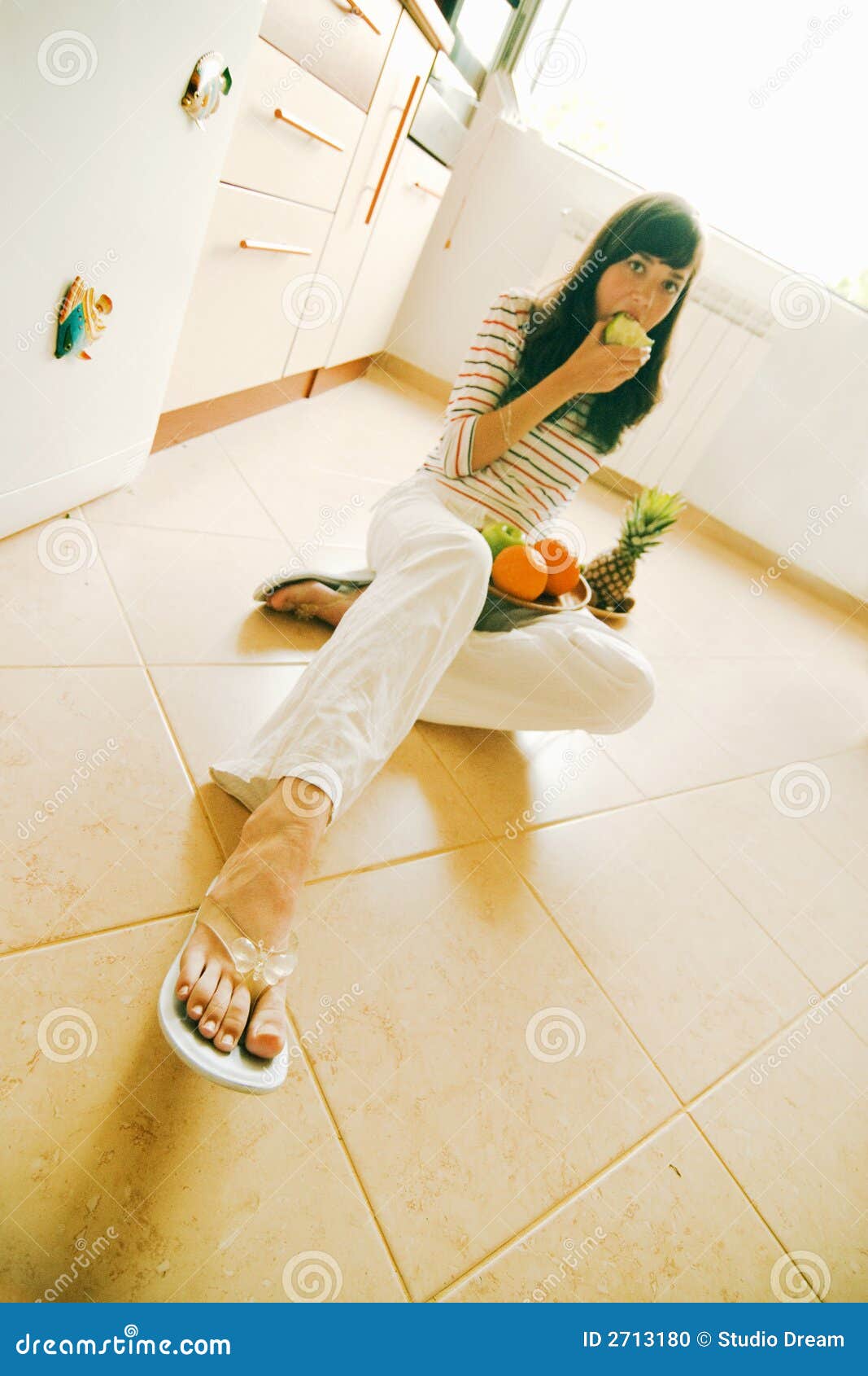 Menina que come a maçã. Menina triguenha que morde de uma maçã verde, sentando-se em um assoalho telhado e com uma cesta da fruta em seu regaço.