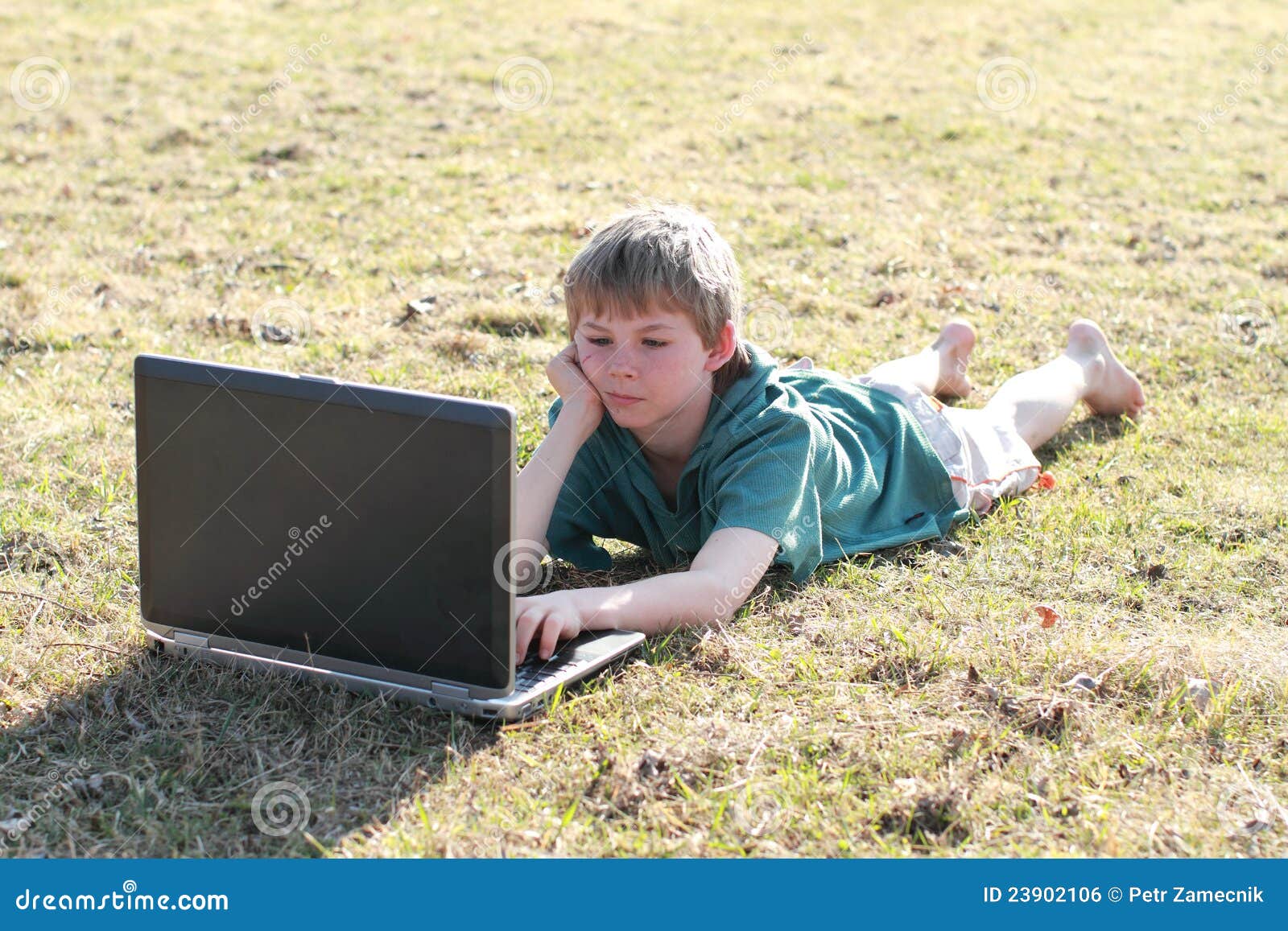 Menino de encontro interessado em um computador. Rapaz pequeno que encontra-se na grama e iterested em um computador.