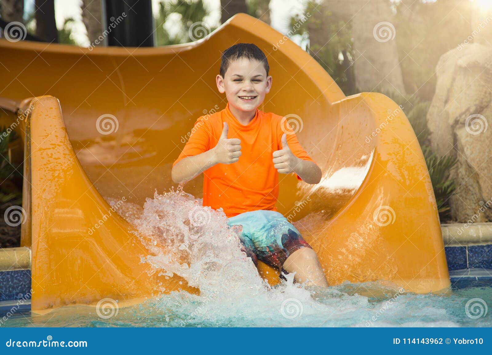 Menino novo de sorriso montando abaixo de mostrar da corrediça de água os polegares acima. Um menino feliz bonito que monta abaixo de uma corrediça de água em um waterpark exterior Dando os polegares até a mostra como está tendo o divertimento