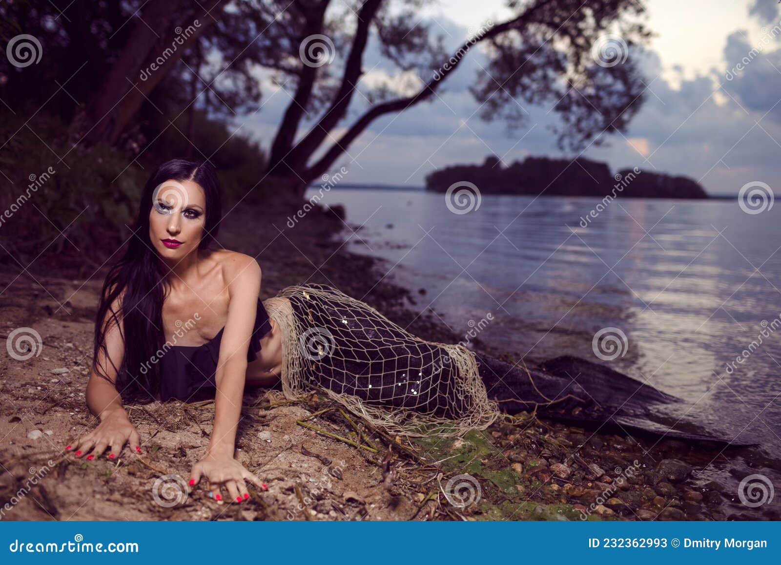 mermaid posing with net near sea on rocks while wearing artistic makeup and black shiny tail covered with colorful spangles posing