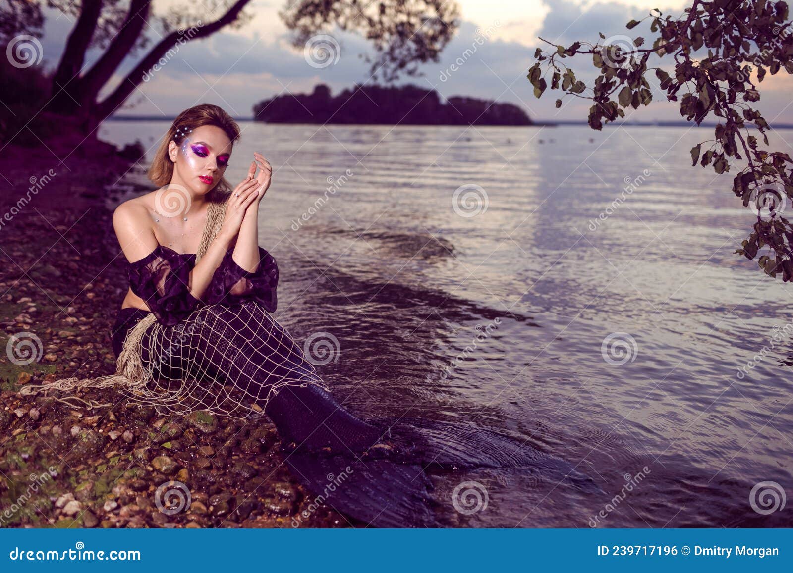 mermaid posing with net near sea on rocks while wearing artistic makeup and facial strassses and black shiny tail covered with