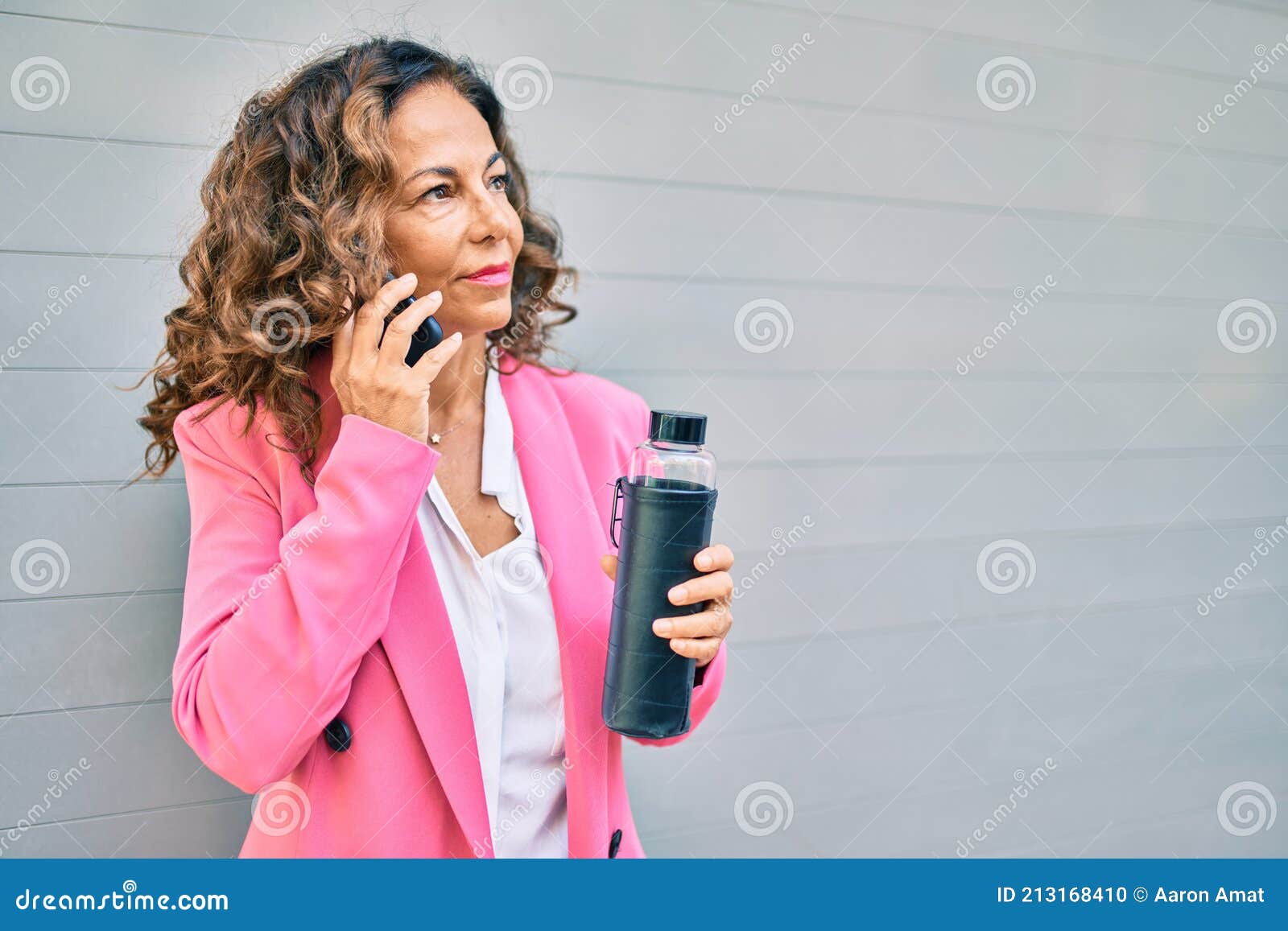 middle age hispanic businesswoman with serious expresison talking on the smartphone and holding bottle of water at the city