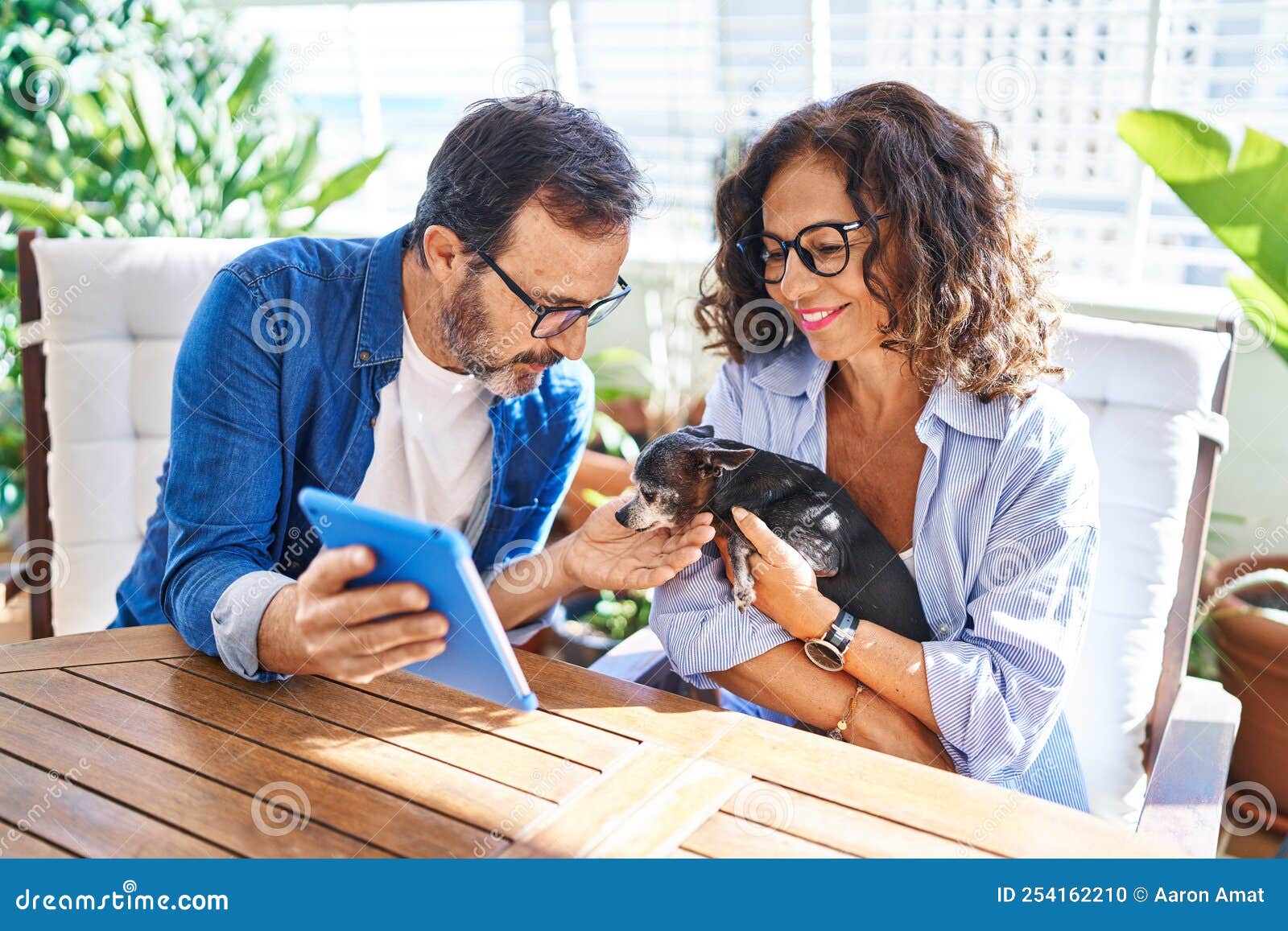 Middle Age Hispanic Couple Using Touchpad Sitting on Table with Dog at Terrace Stock Photo